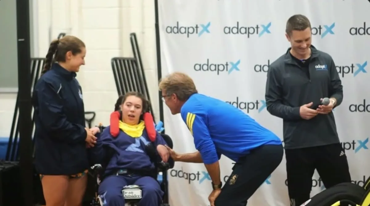 A Hoyt Running Chair presentation in front of an AdaptX backdrop. A young athlete in a wheelchair smiles as a man in a blue jacket kneels to greet her.