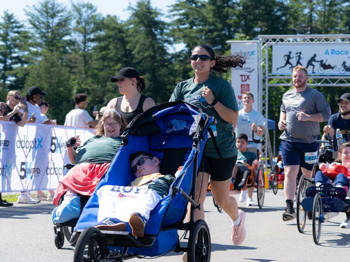 A runner pushing a rider in a blue racing chair past the start banner, other duo teams alongside them