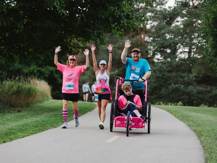 Three people waving on a park path, one of them pushing a rider in a pink racing chair