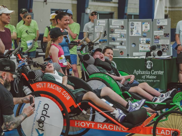 A line of duo teams in orange and green racing chairs waiting to start outside the Aphasia Center of Maine