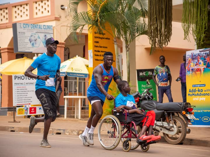 Runners pushing riders in racing wheelchairs along a city street past shopfronts and market umbrellas