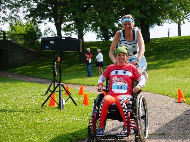 A mother pushing her daughter in a red racing chair along a grass-lined course