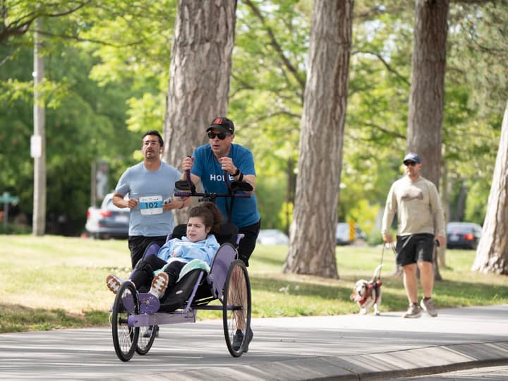 A runner in a cap pushing a rider along a tree-lined park path, another runner beside them