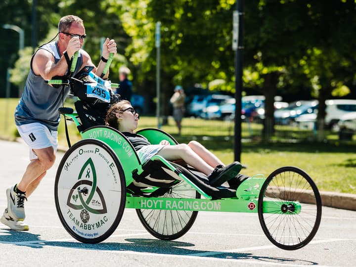 A runner in sunglasses pushing a rider in a green recumbent racing chair along a sunlit road