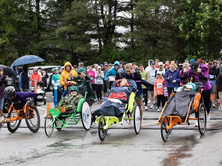 A line of duo teams waiting at a wet start line, the crowd behind them in rain jackets and under an umbrella