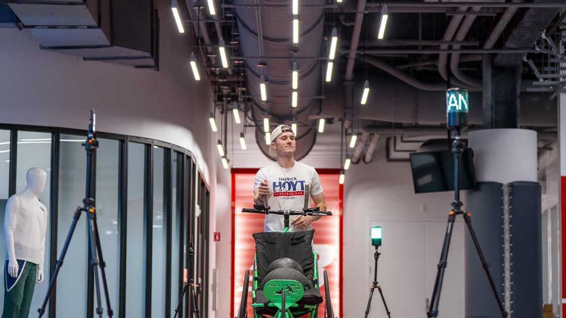 A runner in a Rick Hoyt Research Lab shirt pushing a green running chair along an indoor track at New Balance, flanked by camera tripods recording the run