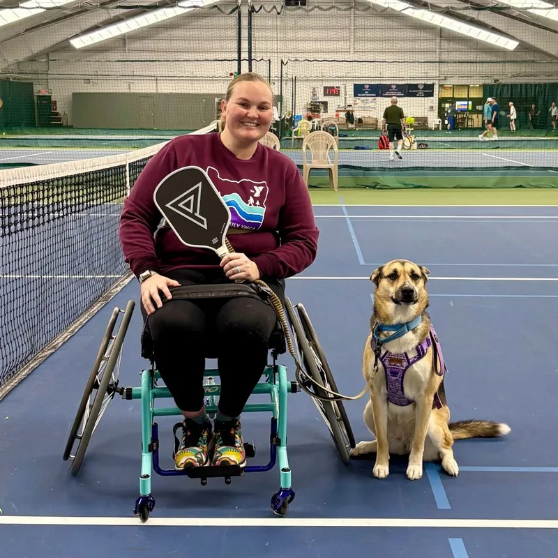 Jess on an indoor court in her sport wheelchair, holding a pickleball paddle, with her service dog sitting beside her