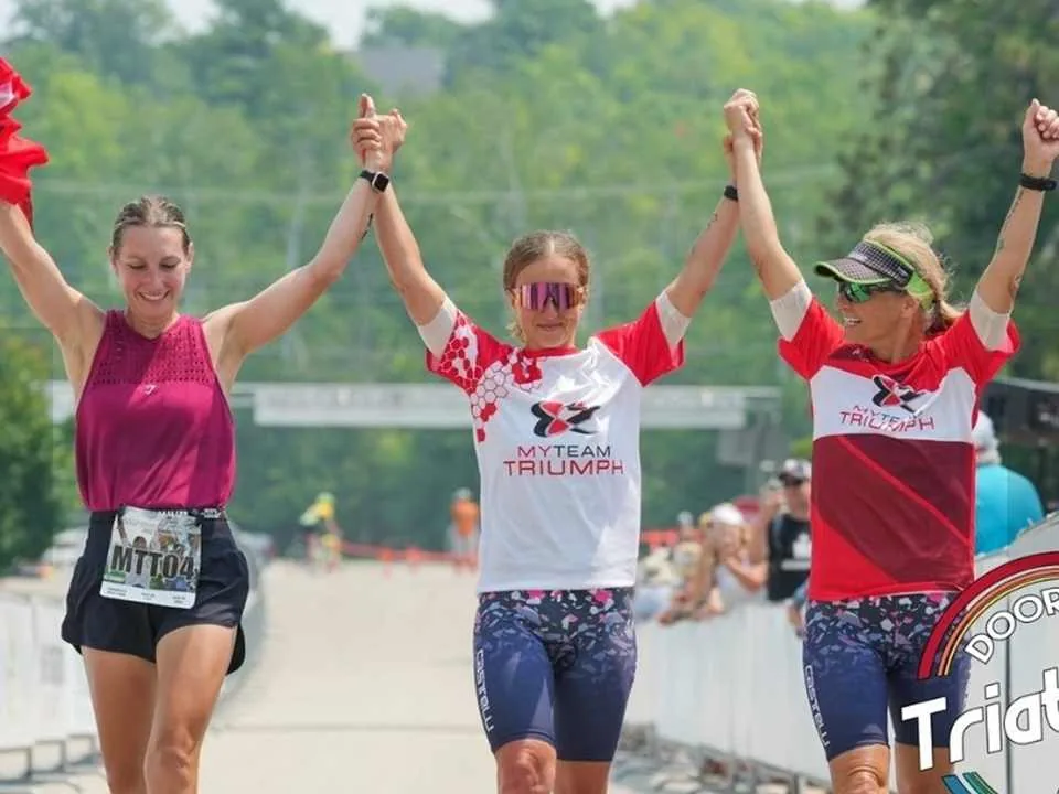 Brenda crossing a triathlon finish line hand in hand with two other athletes, all three with their arms raised