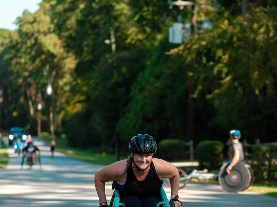 Daria racing on a tree-lined road in a push-rim racing chair, wearing a helmet and racing gloves