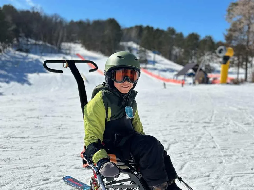 Hudson sitting in a mono-ski on a snowy slope, wearing a helmet and goggles, with a ski lift behind him