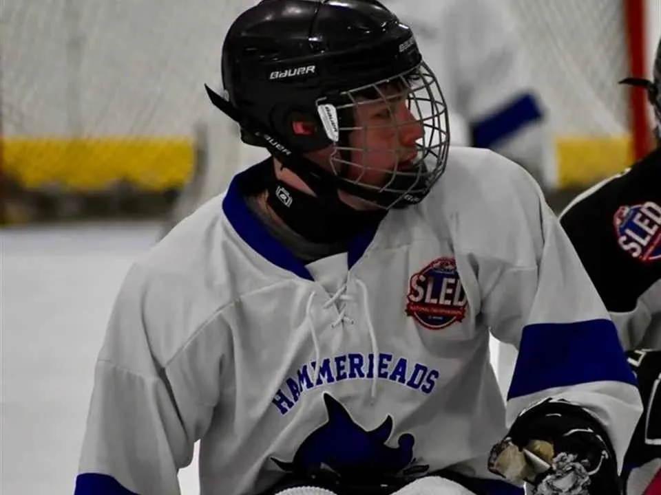 James on the ice in a Hammerheads sled hockey jersey and helmet, seated in his sled holding two sticks