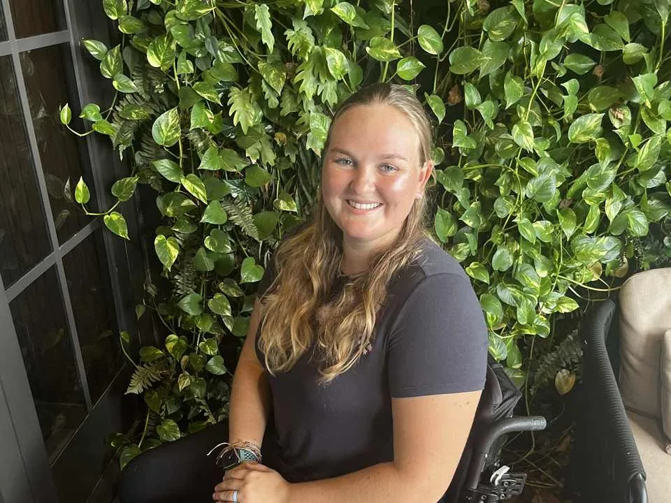 Jess seated in her wheelchair in front of a wall of green foliage, smiling at the camera