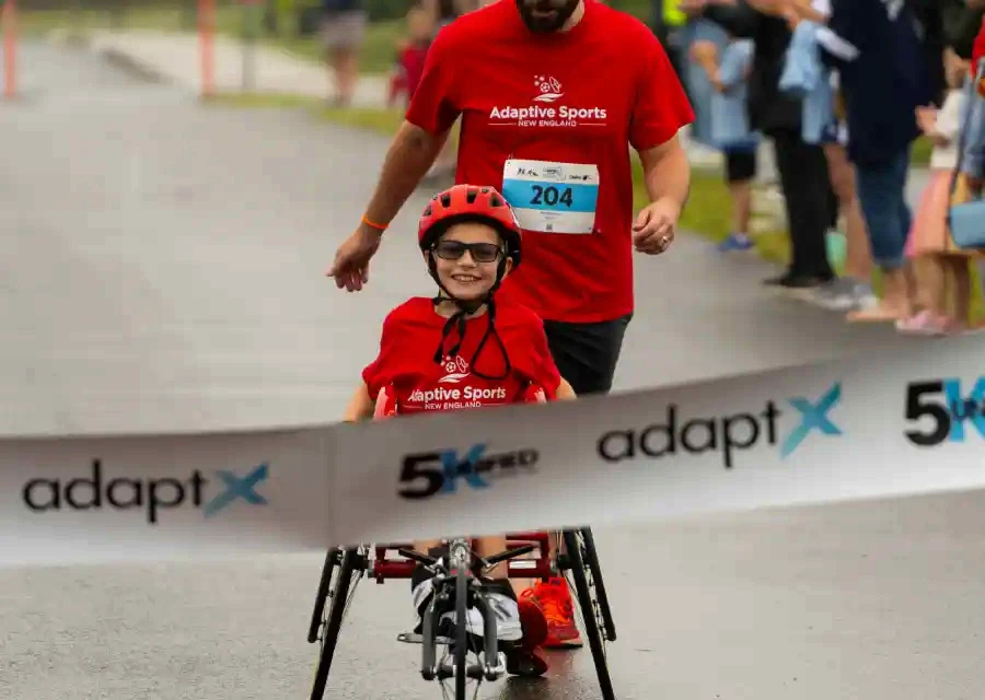 A young athlete in a helmet riding an adaptive bike alongside a running guide near a race finish line