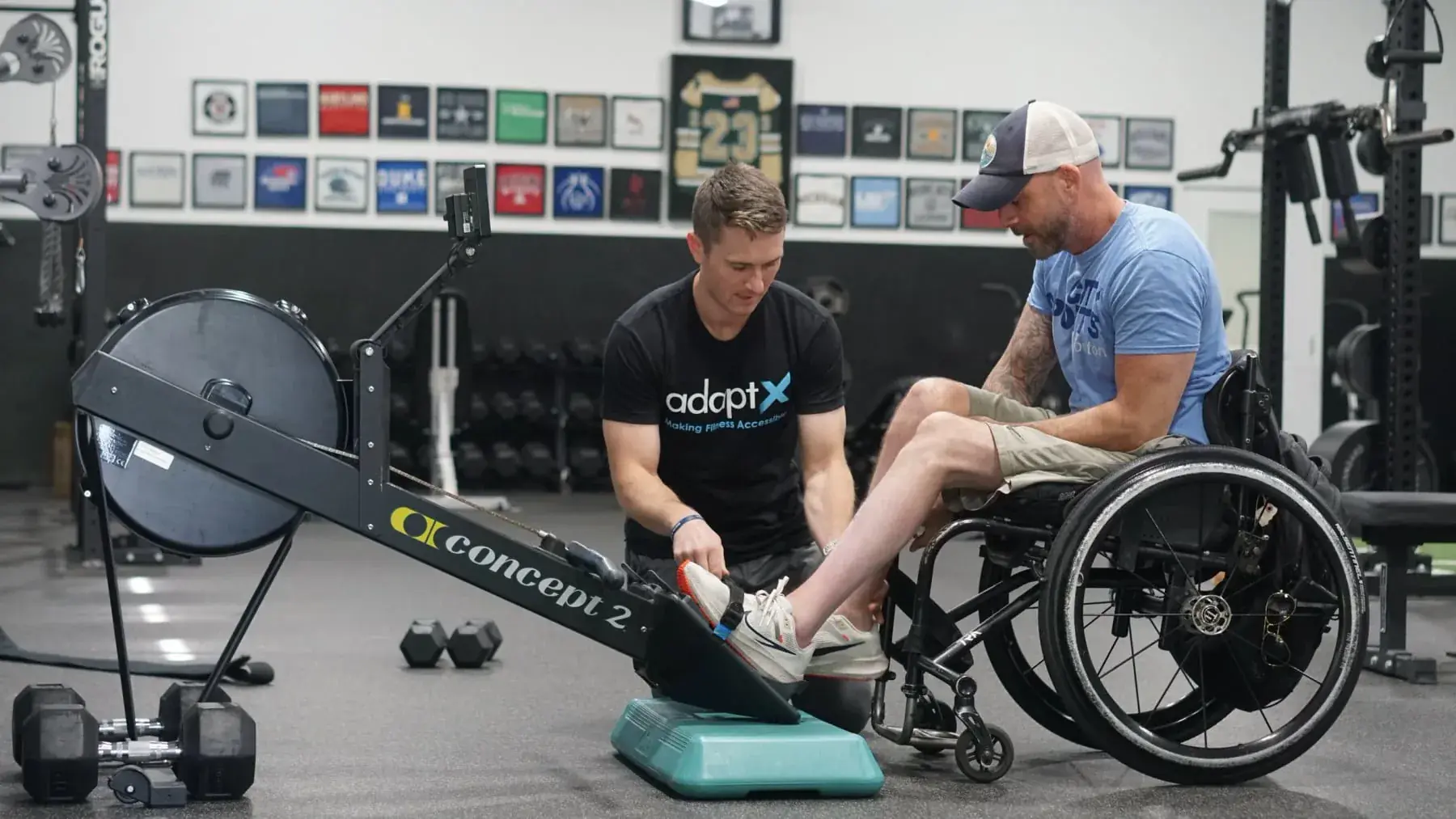 A coach in an AdaptX shirt kneeling beside a rowing machine to help a man in a wheelchair place his foot on the footplate, in a gym hung with framed jerseys