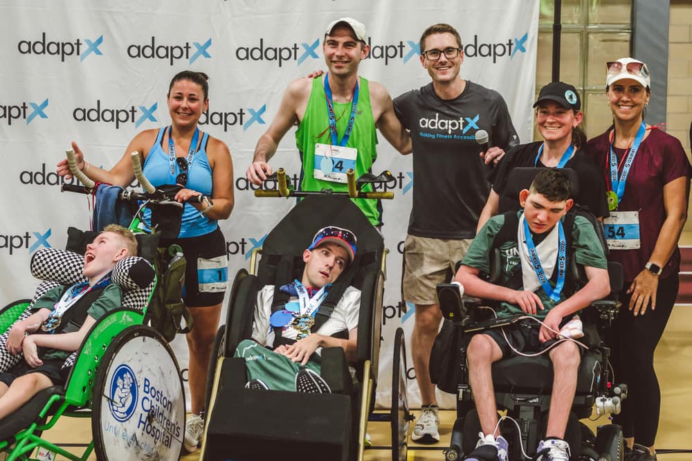 Three duo teams after a race, each athlete in a racing chair beside the runner who pushed them, medals round their necks, in front of a backdrop printed with the AdaptX logo