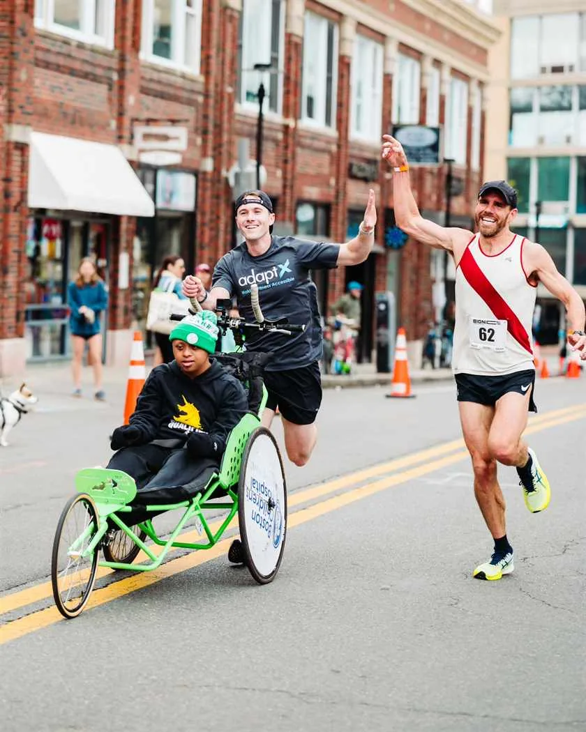 Brendan Aylward racing with Jacob in a green running chair, high-fiving another runner as they pass on a city street