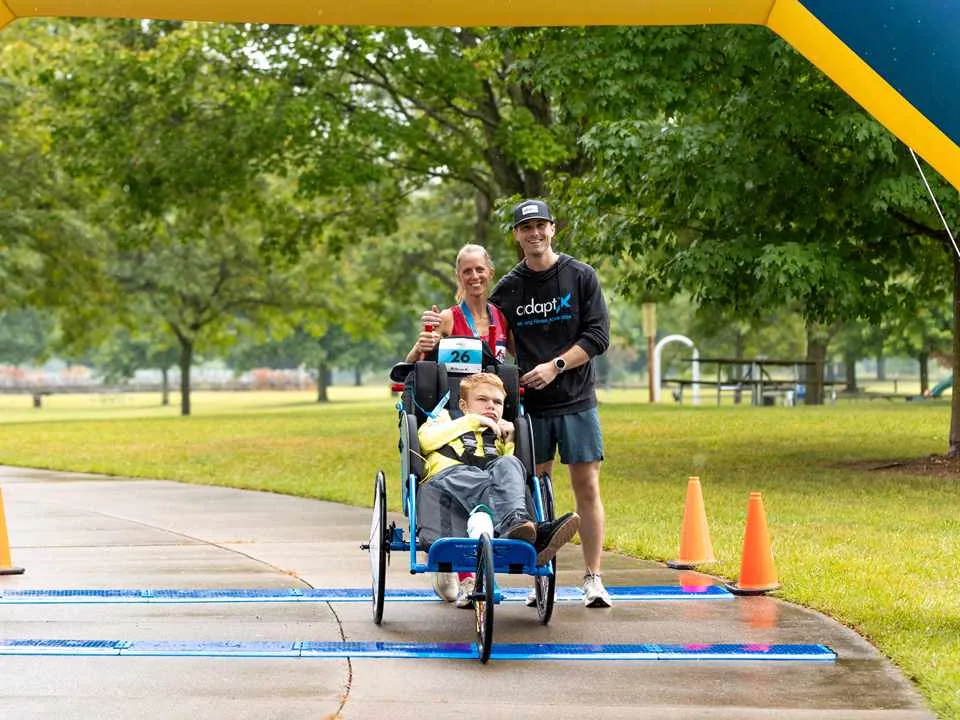 Austin at the finish line of the Dexter, Michigan Unified 5K in a blue running chair, with Sandy and Brendan Aylward beside him