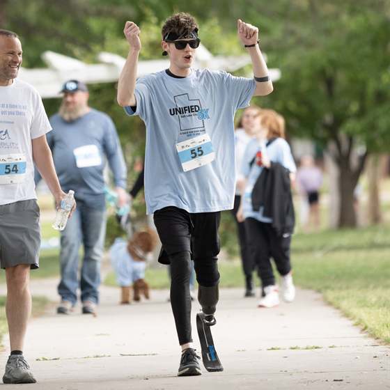 A young runner on a running blade, both arms raised, wearing a Unified 5k race bib on a tree-lined course