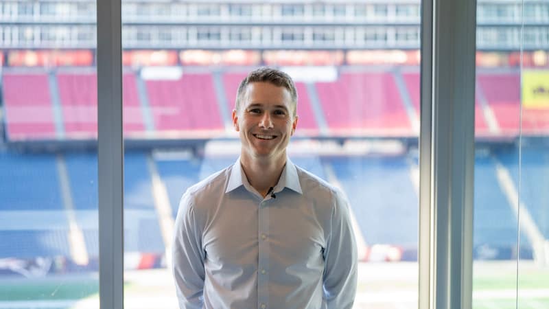 Brendan Aylward photographed in front of a stadium window, tiered seating and the pitch behind him.