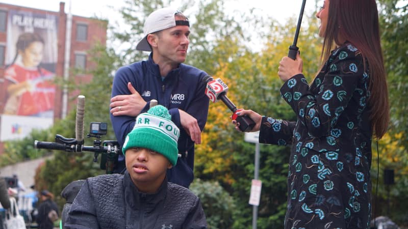 A man in a cap being interviewed by a reporter holding a microphone, with a person in a green beanie seated in front of them.