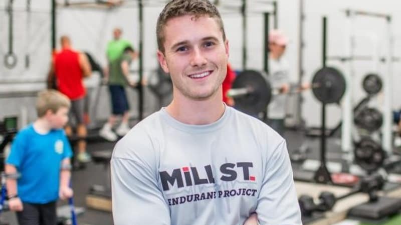 A young man in a training shirt standing with his arms crossed in a gym, smiling, with others working out behind him.