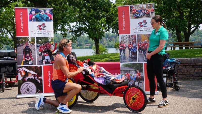 A man crouched beside a red racing chair with a young rider in it, in front of banners of race photographs, with a woman standing to the right.