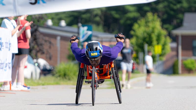 An athlete in a racing chair coming head-on toward the camera with both arms raised, spectators lining the road behind.