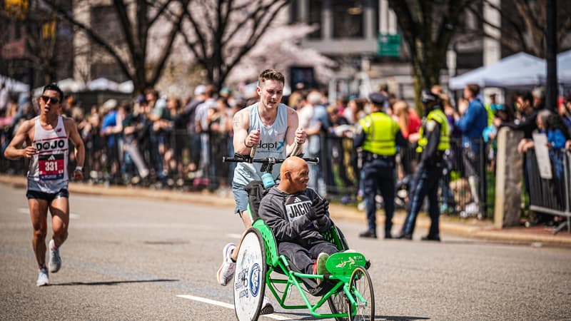 A runner pushing a green racing chair along a city street lined with spectators and police barriers, another runner alongside.