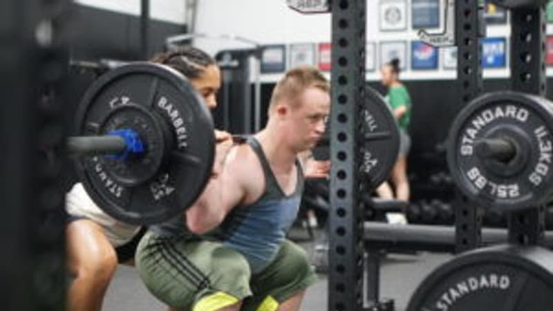 Two people squatting with loaded barbells side by side in a gym rack area.