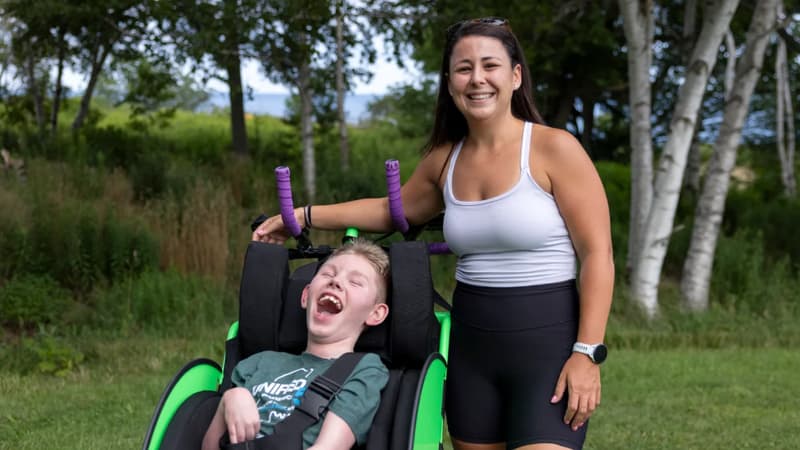 A young person seated in a green running chair, head tipped back and laughing, with a woman standing beside them on grass.