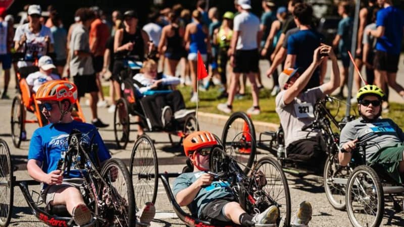 A row of young athletes in handcycles and racing chairs lined up on a road with a large field of runners massed behind them.