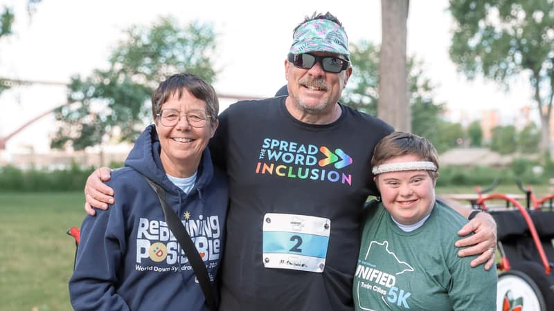 Three people standing arm in arm and smiling: an older woman, a man wearing a race number, and a young man with Down syndrome.
