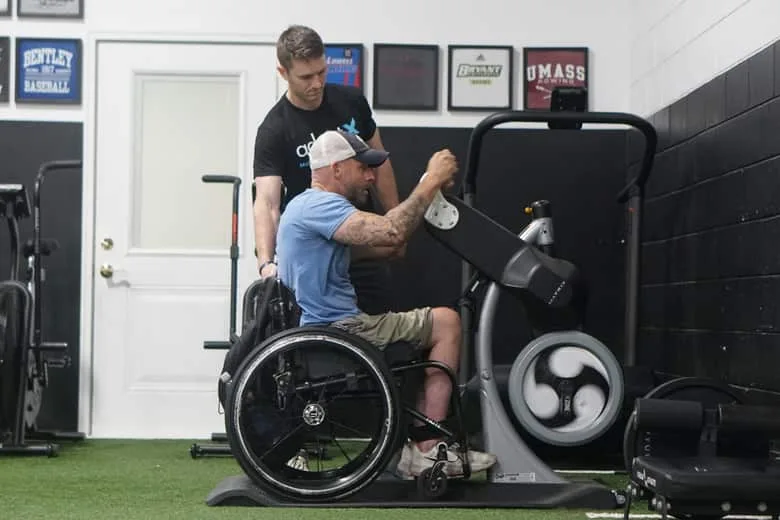 A coach standing over a wheelchair user working at an arm cycle.