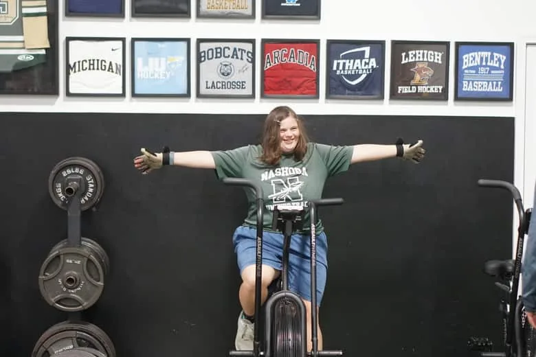 A young woman on an air bike throwing both arms wide, grinning, at the end of an effort.