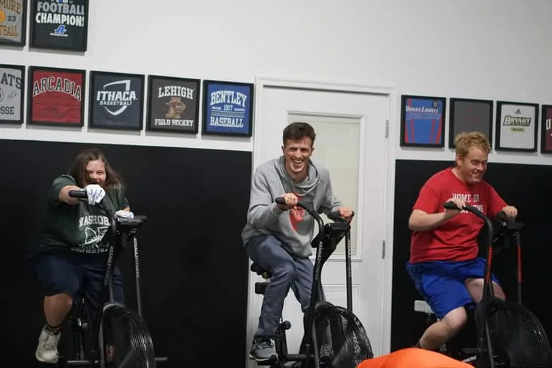 Three athletes riding air bikes side by side along the gym wall.