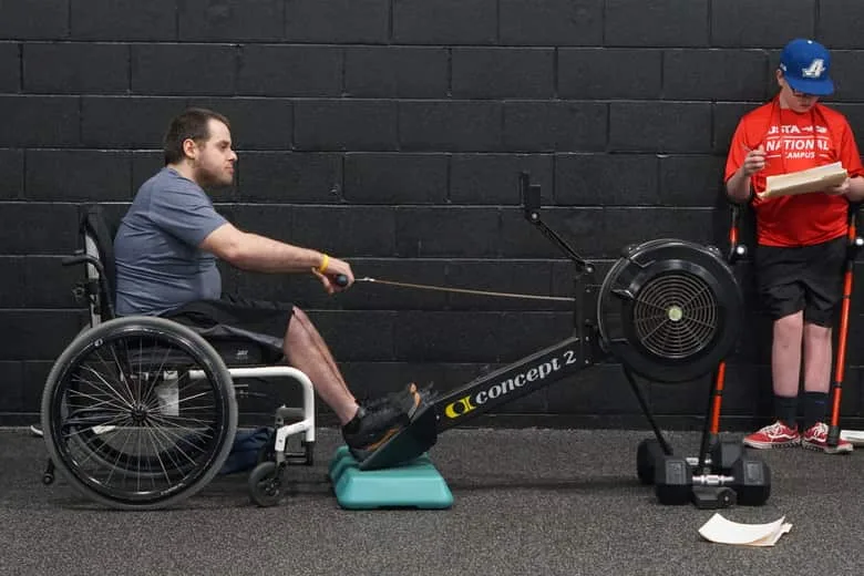 A wheelchair user rowing on an adapted erg while another athlete waits nearby on forearm crutches.