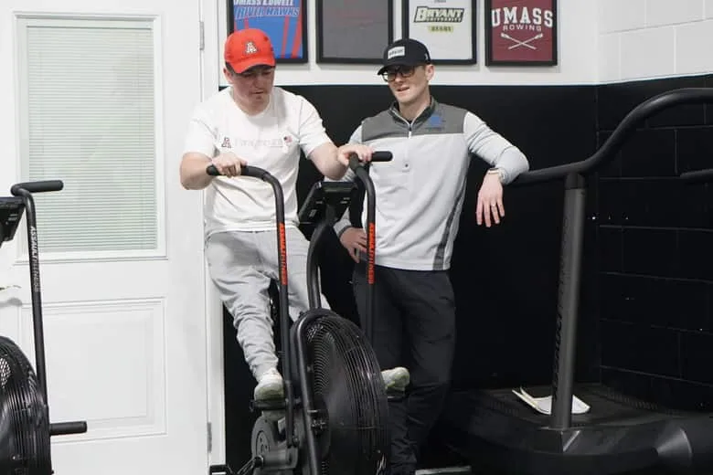 Two athletes at the air bikes, one riding and one resting on the handlebars beside him.