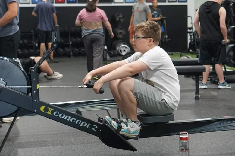 A young athlete rowing on an erg, mid-drive, with the rest of the session going on behind him.