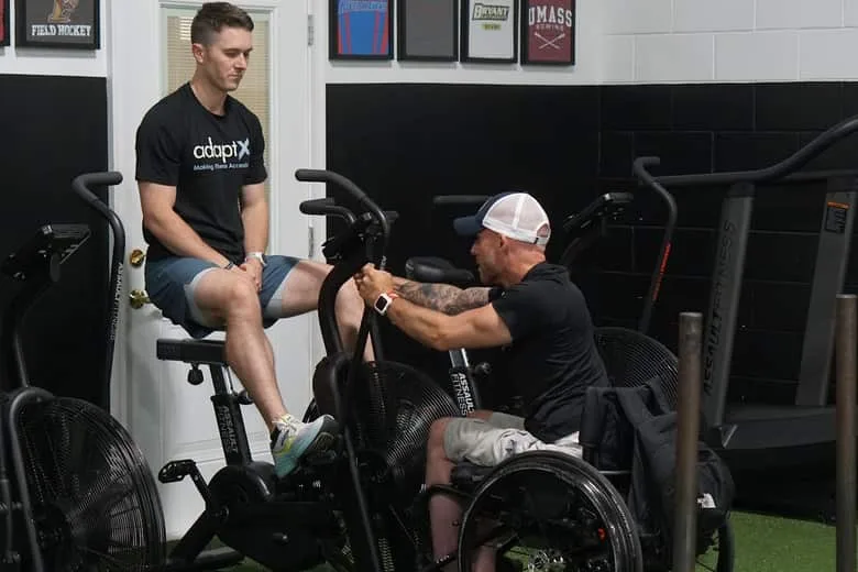 A coach on an air bike beside an athlete in a wheelchair setting up at the machine next to him.