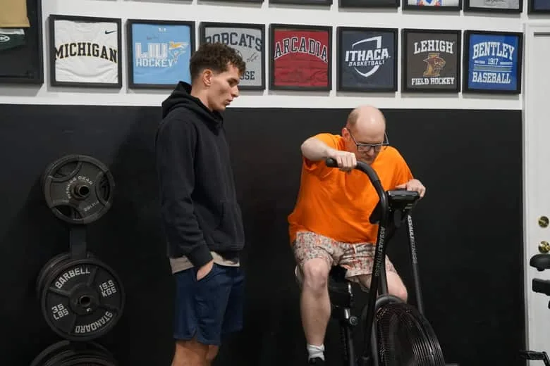 An athlete riding an air bike while a training partner stands alongside.