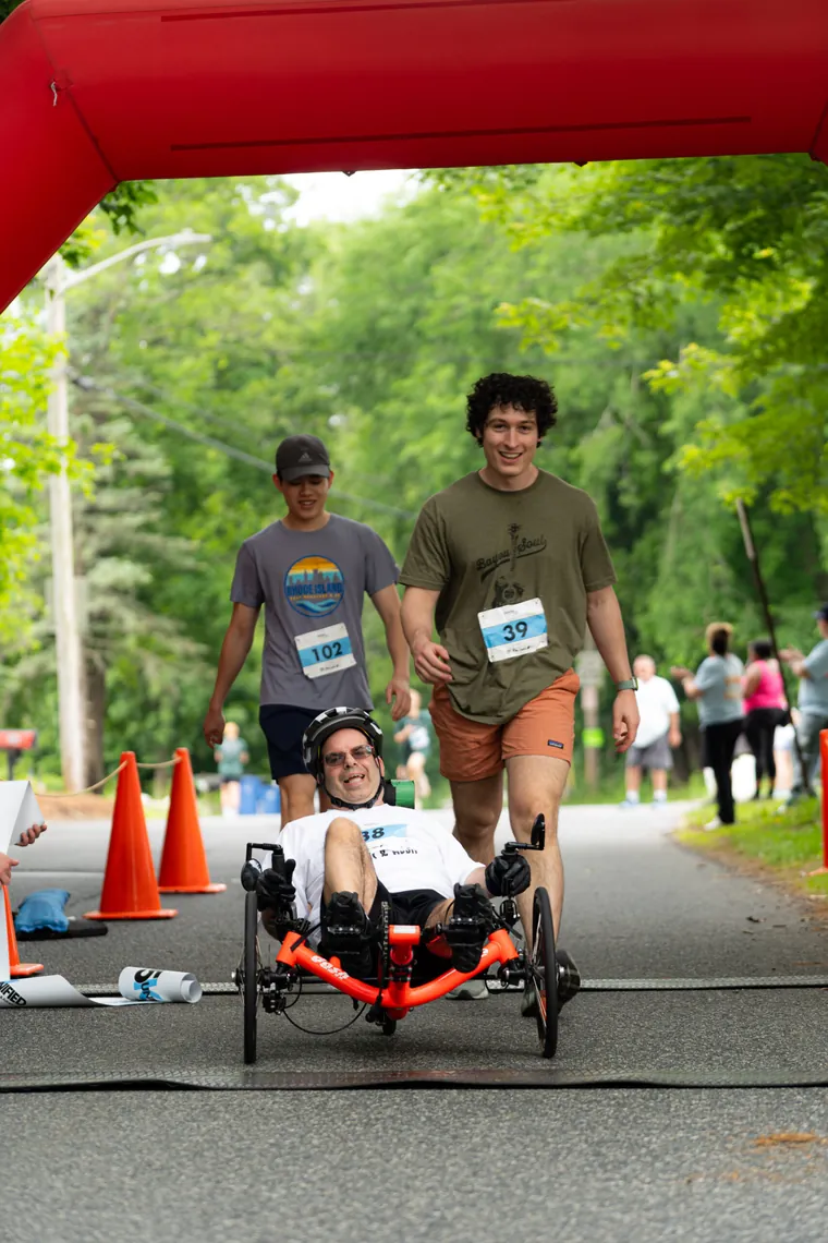 Two runners pushing a handcycle rider under the start arch