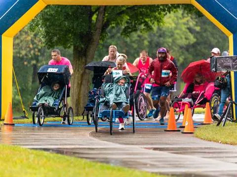 Duo teams racing through a blue and yellow inflatable arch in the rain, the riders wearing ponchos in their running chairs