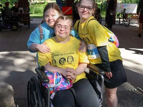 Three participants in yellow race shirts together, one seated in a wheelchair with the other two leaning in beside her
