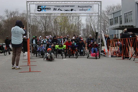 Adaptive athletes lined up in racing chairs and handcycles under the Unified 5K start arch